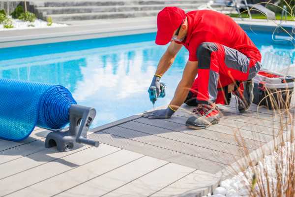 Worker in red uniform performing pool maintenance, using a drill on decking near a swimming pool, with rolled pool cover and maintenance tools visible. Worker in red uniform performing pool maintenance, using a drill on decking near a swimming pool, with rolled pool cover and maintenance tools visible.
