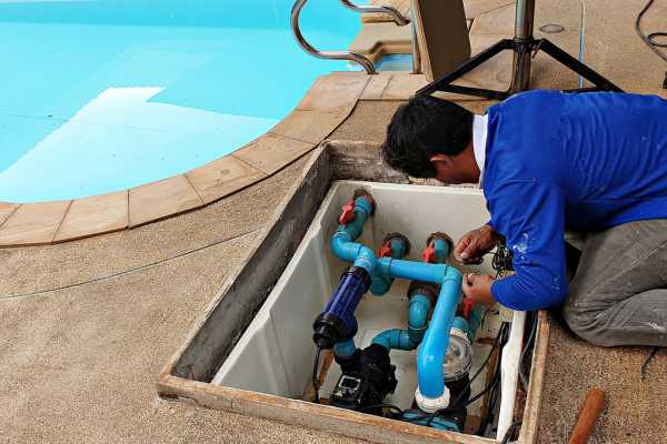 Pool maintenance technician inspecting plumbing system, ensuring efficiency for storm season, with swimming pool in background.